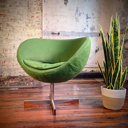 Green egg-shaped chair in a room with a brick wall and a potted plant.