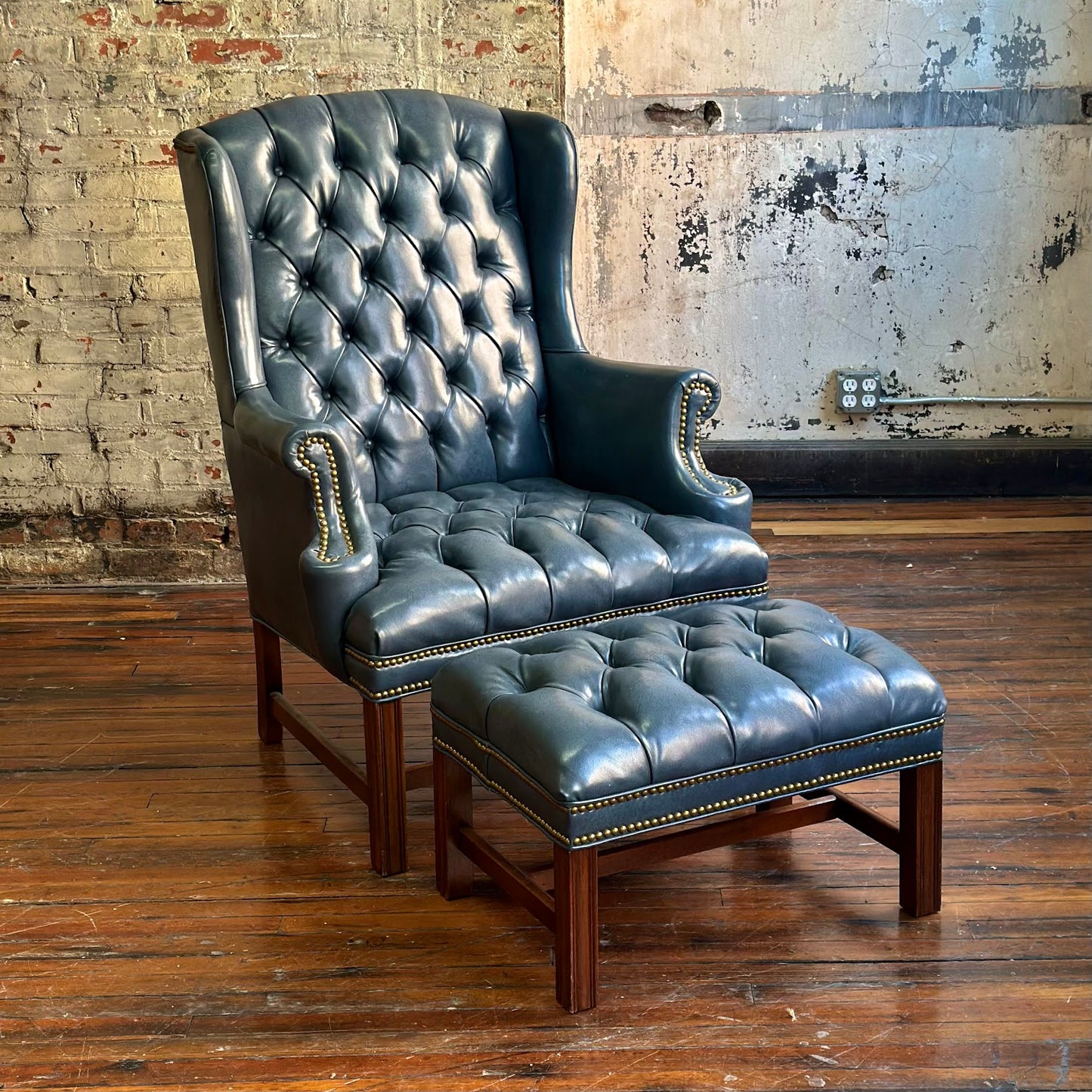 Blue leather armchair with matching ottoman on a wooden floor against a brick wall.