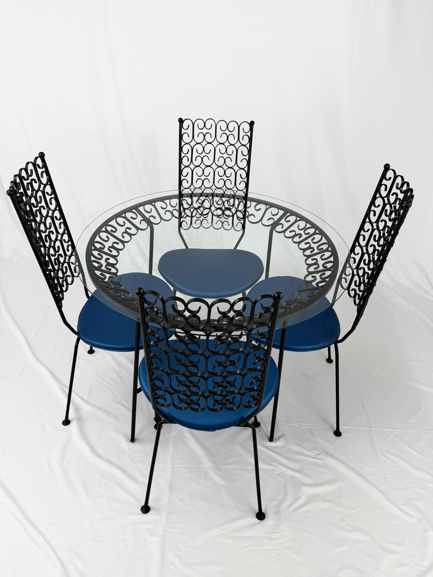 Wrought Iron dining set with four blue cushioned chairs and a round glass top table on a white background