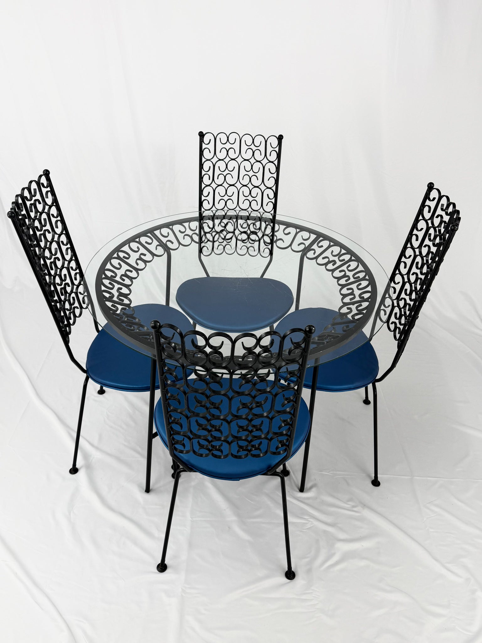 Wrought Iron dining set with four blue cushioned chairs and a round glass top table on a white background