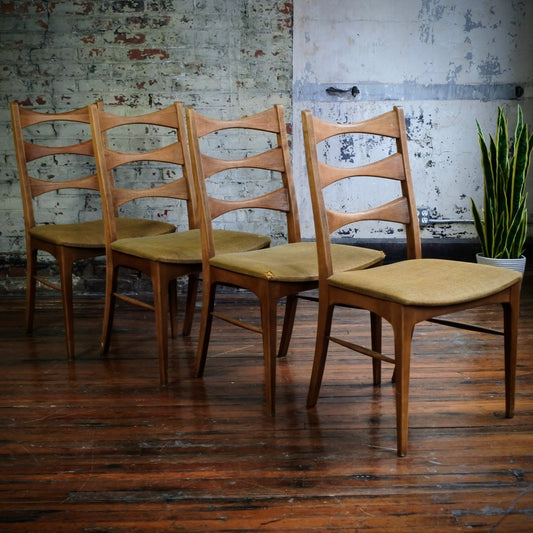 Four wooden chairs with beige cushions against a textured brick wall.