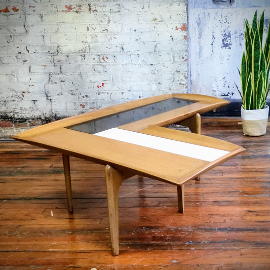 Wooden boomerang coffee table with tile top on a wooden floor with a plant in the background.