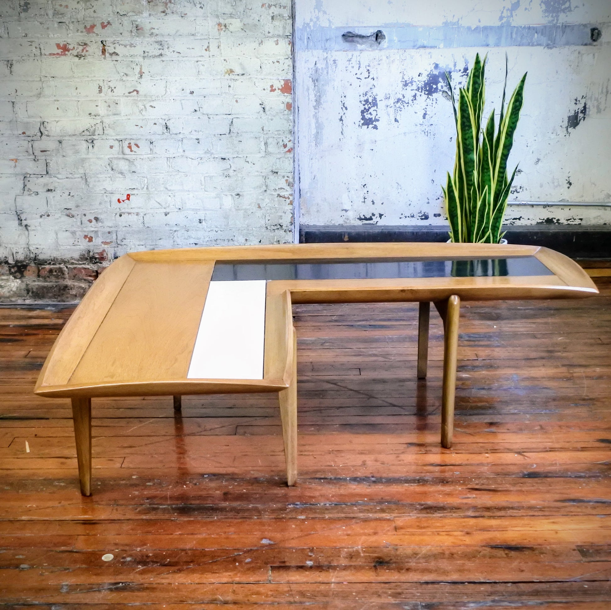 Wooden boomerang coffee table with tile top on a wooden floor with a plant in the background.