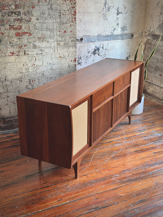 A wooden audio console with sliding doors and control panel, placed on a wooden floor against a brick wall background.