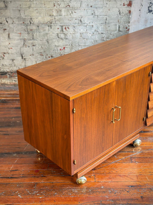 A brown wooden stereo console on casters, featuring multiple drawers and cabinet doors, positioned against a brick wall with a plant beside it.