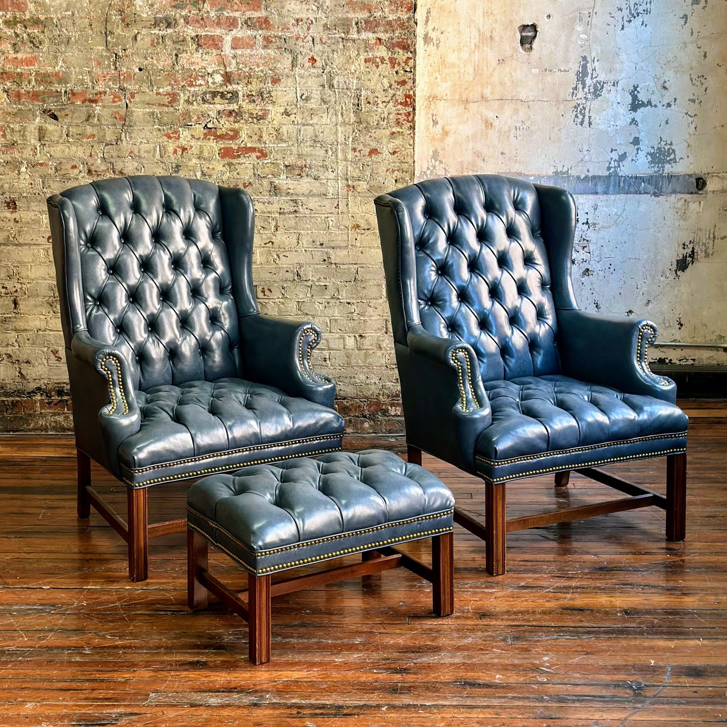 Two blue leather armchairs with matching ottoman against a brick wall.