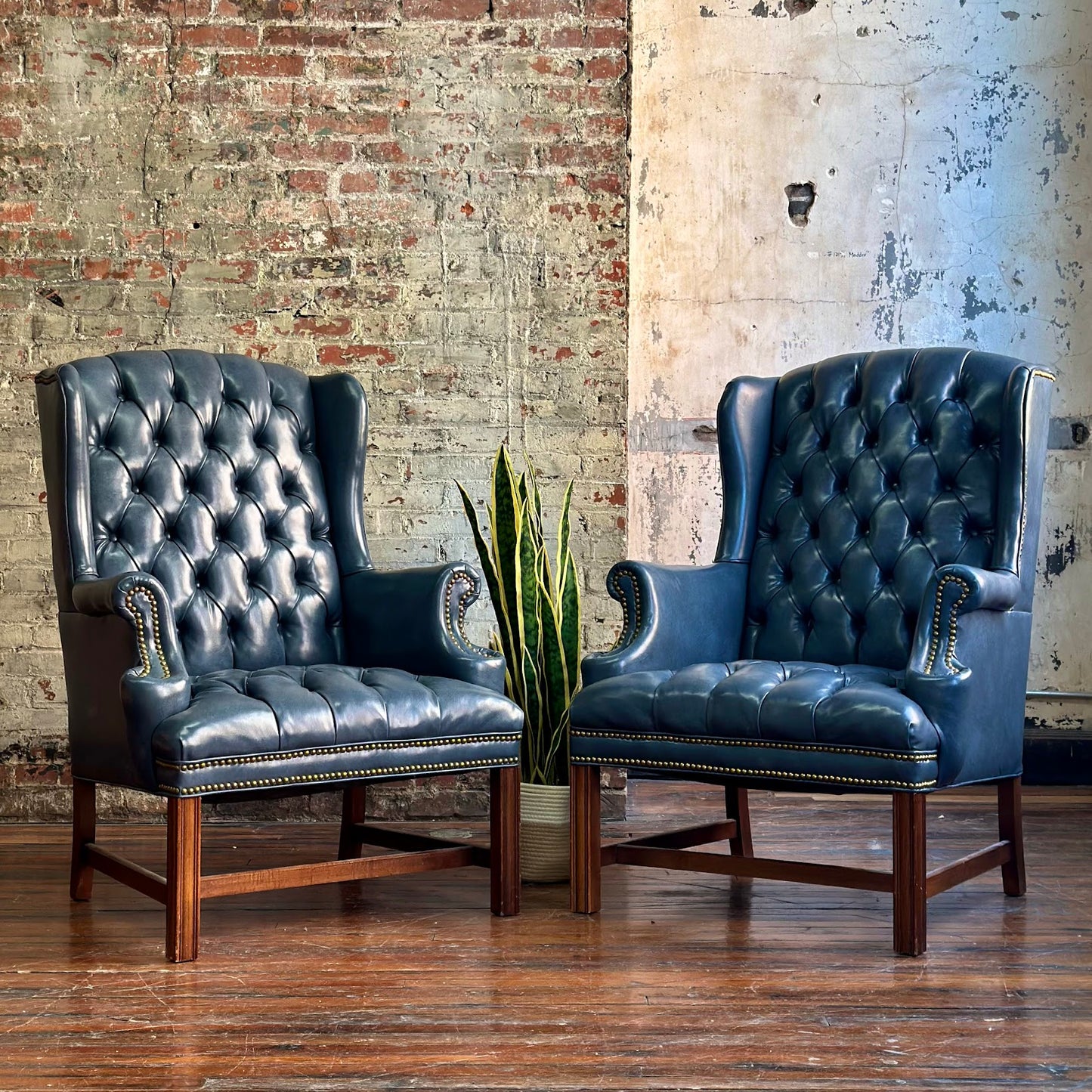 Two blue leather armchairs with wooden legs in front of a brick wall.